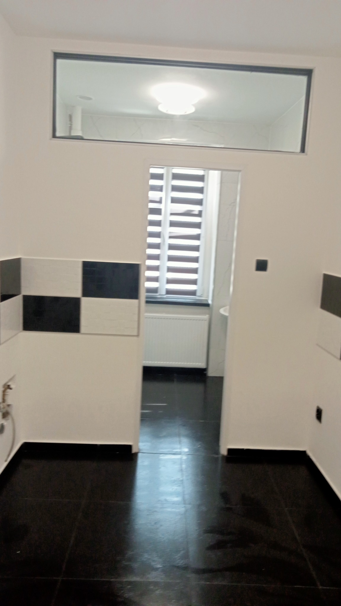 Wnętrze łazienki po remoncie, widoczne czarne płytki podłogowe, fragment ściany z biało-czarną szachownicą z płytek, okno z żaluzjami oraz zamontowane oświetlenie sufitowe.