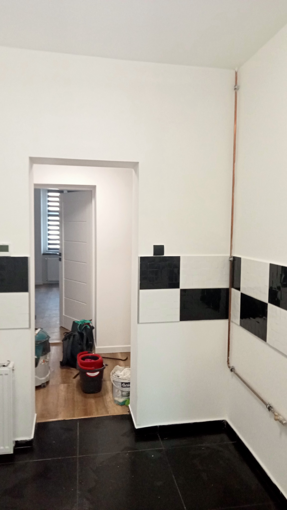 Wnętrze po remoncie: białe ściany z czarno-białą szachownicą z płytek, czarna podłoga, widoczne rury miedziane, otwarte drzwi do innego pomieszczenia z widocznym sprzętem sprzątającym i wiadrem...
