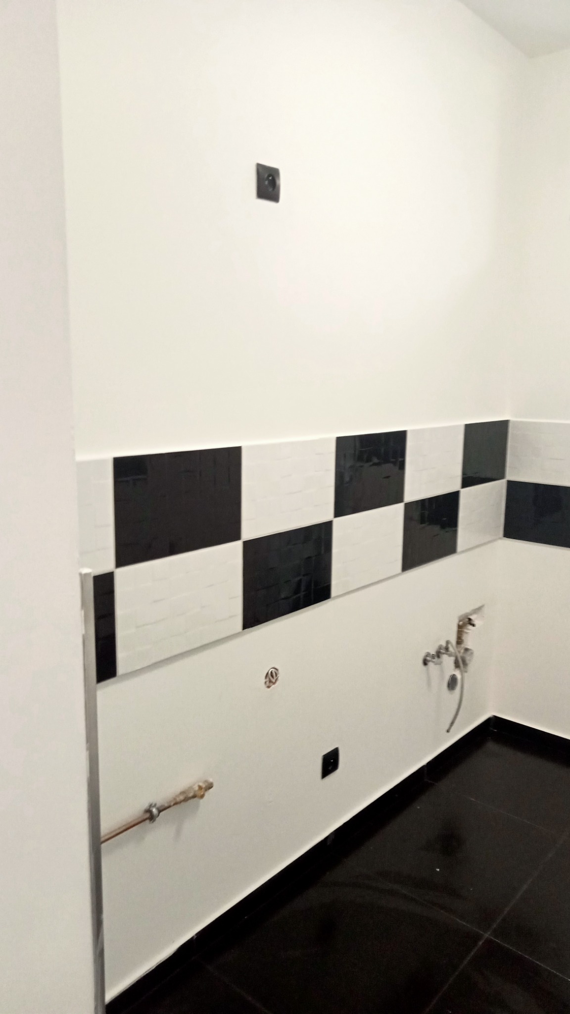 Niedokończona łazienka z czarno-białą szachownicą z płytek na ścianie, widoczne wystające rury i gniazdko elektryczne, czarne płytki podłogowe.