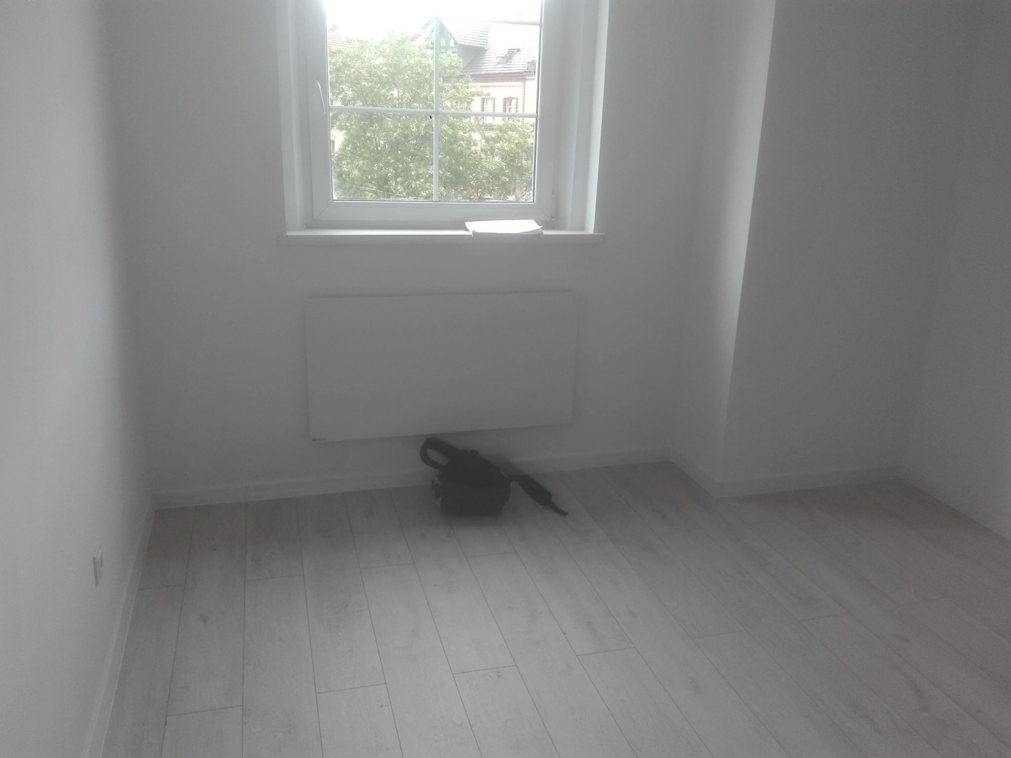 Pusty pokój z świeżo pomalowanymi białymi ścianami, jasną podłogą imitującą drewno i oknem z widokiem na drzewa. Na parapecie leżą białe kartki. Pod grzejnikiem leży czarna torba.