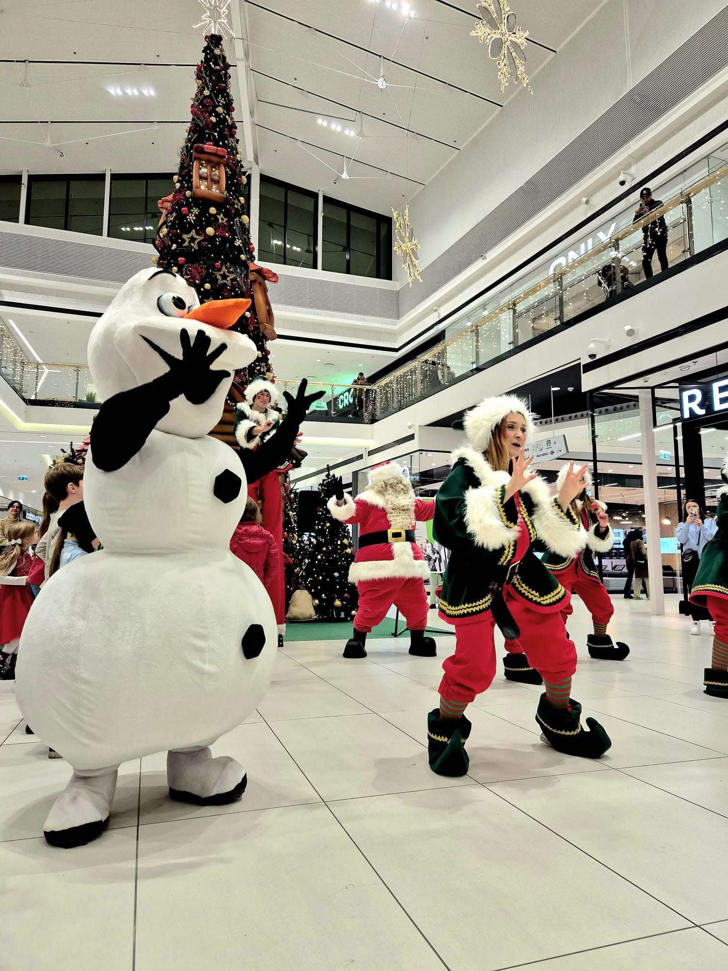 Występ postaci w kostiumach: bałwan Olaf, Mikołaj i elfy w centrum handlowym na tle choinki.