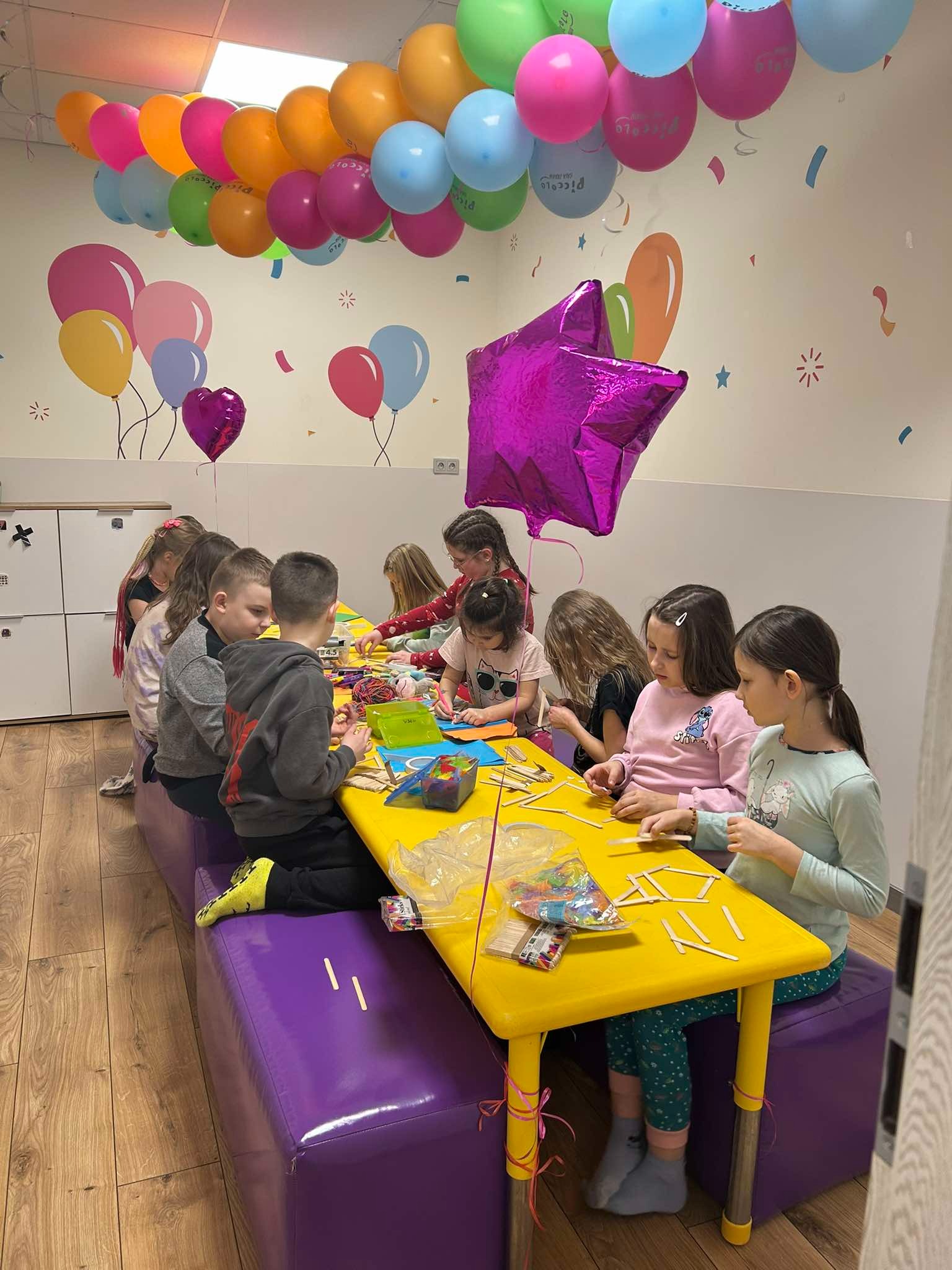 Grupa dzieci siedzi przy żółtym stole, wykonując prace plastyczne z patyczków i kolorowego papieru, w tle dekoracje balonowe i ścianka z motywami balonów.