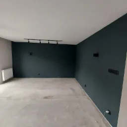 Pusty pokój po malowaniu ścian na ciemny odcień zieleni, widoczna szyna z reflektorami na suficie, gniazdka elektryczne i grzejnik. Podłoga surowa, przygotowana do dalszych prac.