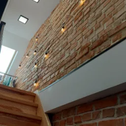 Schody drewniane prowadzące w górę, ściana z czerwonej cegły z dekoracyjnymi żarówkami, biały sufit z kwadratowymi lampami, widok z dołu.