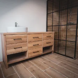 Jasna drewniana szafka łazienkowa z umywalką nablatową i czarnymi uchwytami szuflad, w tle kabina prysznicowa z czarnymi profilami, płytki imitujące drewno na podłodze i ścianie.