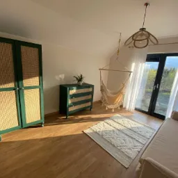 Jasne wnętrze pokoju z zielonymi meblami z rattanowymi frontami: szafa i komoda, hamak fotel wiszący przy oknie, beżowa sofa i dywan na drewnianej podłodze.