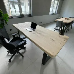 Jasny drewniany blat biurka na czarnych, trapezowych nogach w nowoczesnym biurze, z laptopem, krzesłem biurowym i widokiem na rusztowanie za oknem.