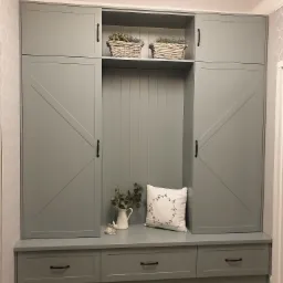 Szara zabudowa meblowa w przedpokoju z siedziskiem i trzema szufladami, górne szafki z dekoracyjnymi drzwiami przesuwnymi w stylu rustykalnym, na półce wiklinowe koszyki z roślinami, na siedzisku...