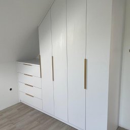 Promonta 2 Marcin Staniszewski - Biała szafa wnękowa z szufladami i złotymi uchwytami dopasowana do skosu poddasza, widoczna na tle jasnych paneli podłogowych.