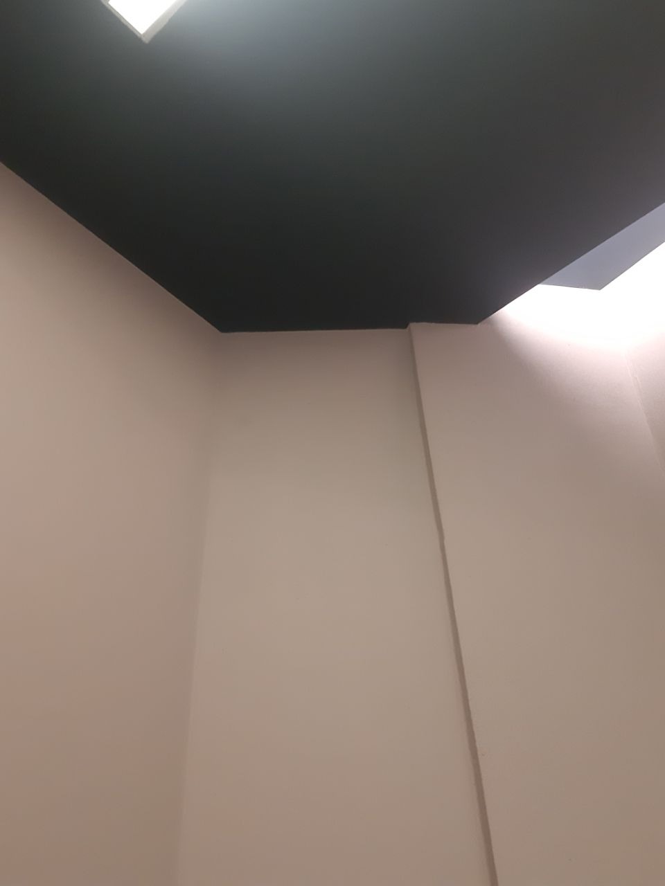 Wnętrze pomieszczenia z pomalowanym na czarno fragmentem sufitu i jasnymi ścianami, widoczne narożniki i oświetlenie sufitowe.