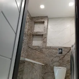 Wykończona łazienka z marmurowymi płytkami, podświetlaną wanną, wiszącą toaletą i pralką, widok zza częściowo otwartych drzwi.