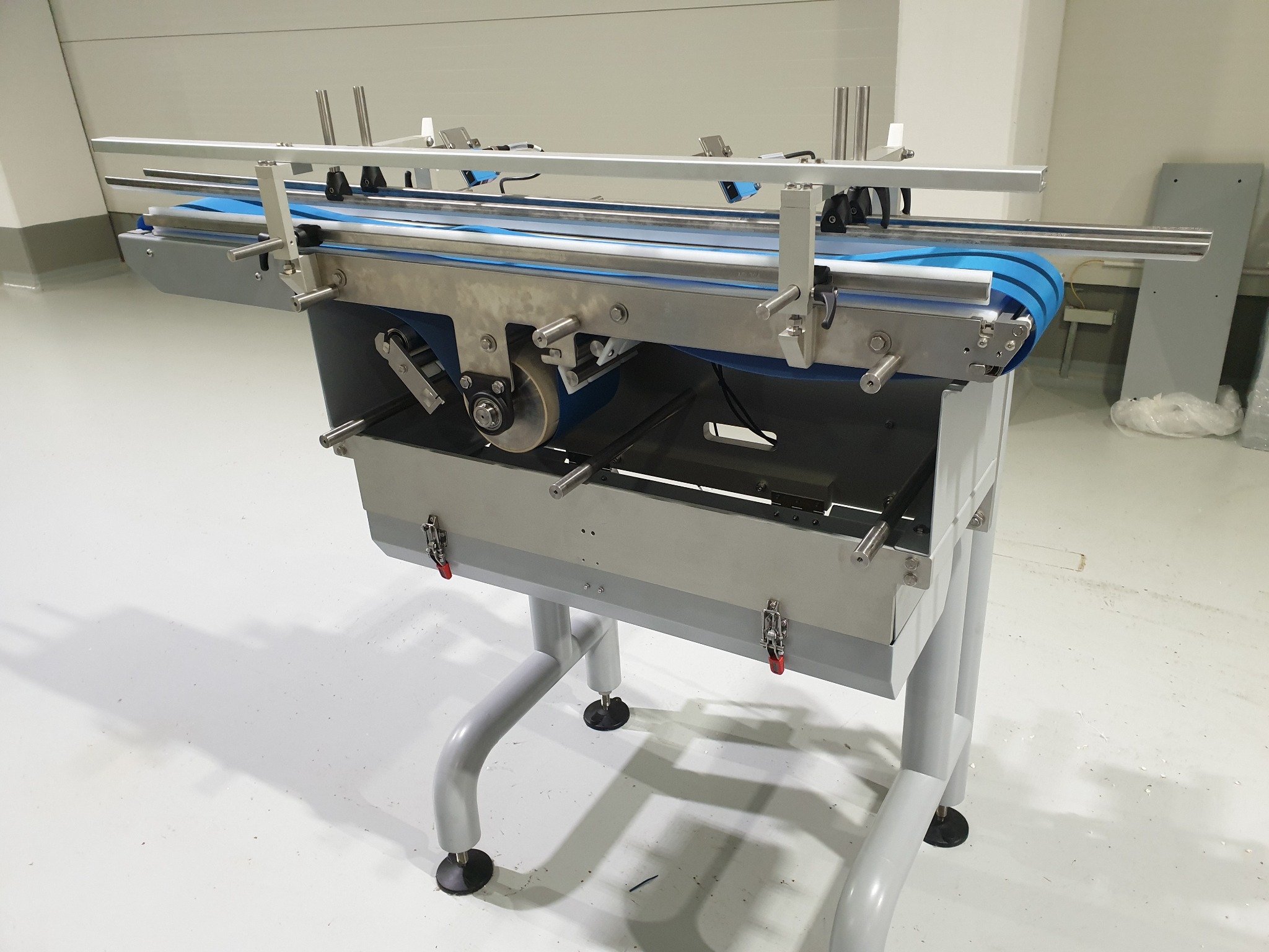 Szczegółowe ujęcie fragmentu linii produkcyjnej z niebieskim taśmociągiem, metalową konstrukcją i regulowanymi nóżkami w sterylnym, białym pomieszczeniu.