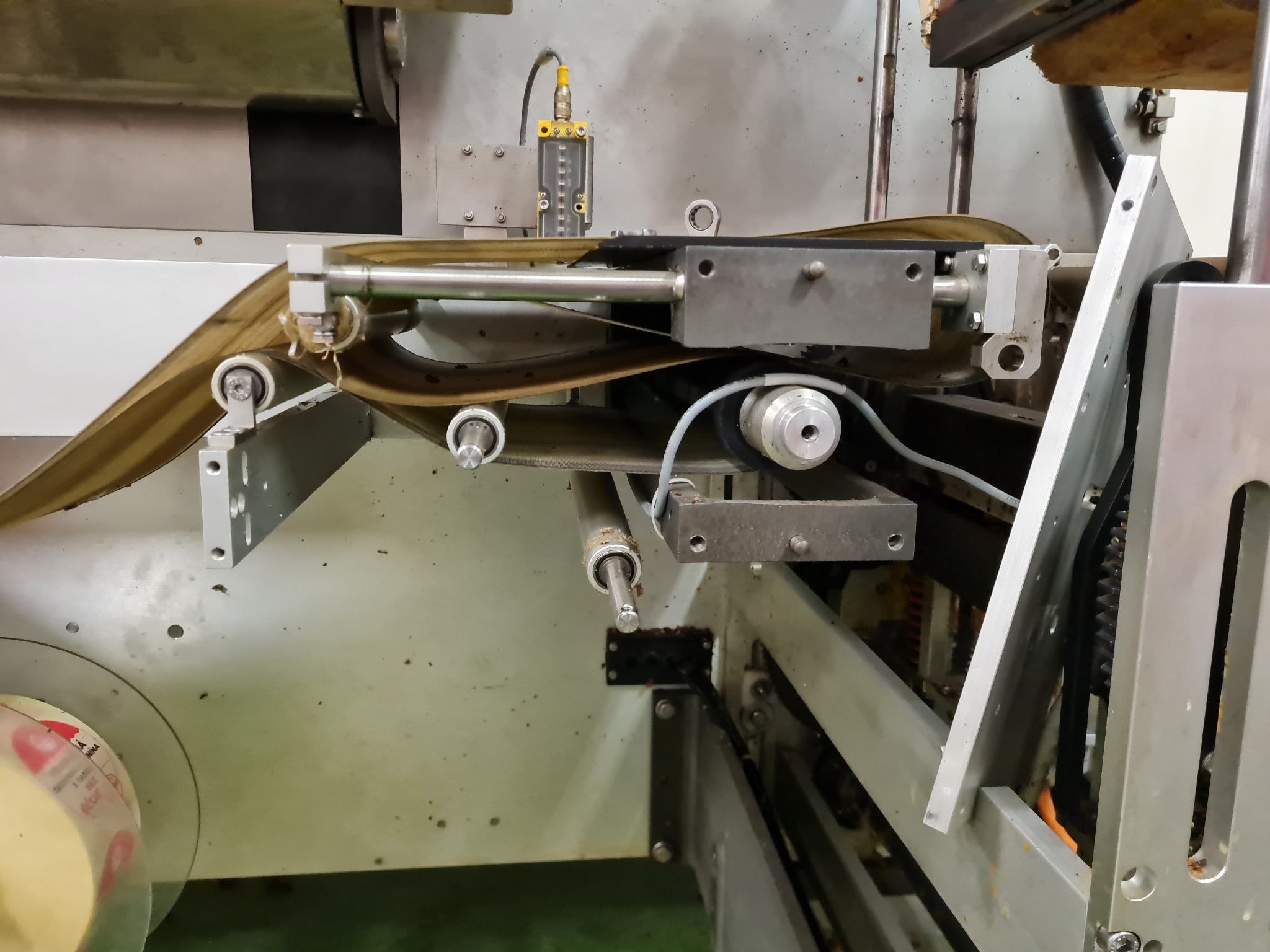 Fragment maszyny przemysłowej z systemem rolek prowadzących taśmę, widoczny element sterujący z rurką pomiarową, całość w odcieniach szarości i beżu.