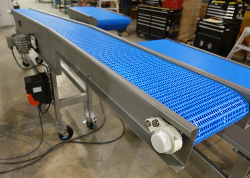 Szara taśma transportowa z niebieską, ażurową powierzchnią roboczą, zamontowana na kółkach z widocznym silnikiem elektrycznym i skrzynką sterowniczą.