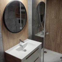 Nowoczesna łazienka z umywalką nablatową, okrągłym lustrem z czarną ramą, kabiną prysznicową typu walk-in i drewnianymi panelami ściennymi.