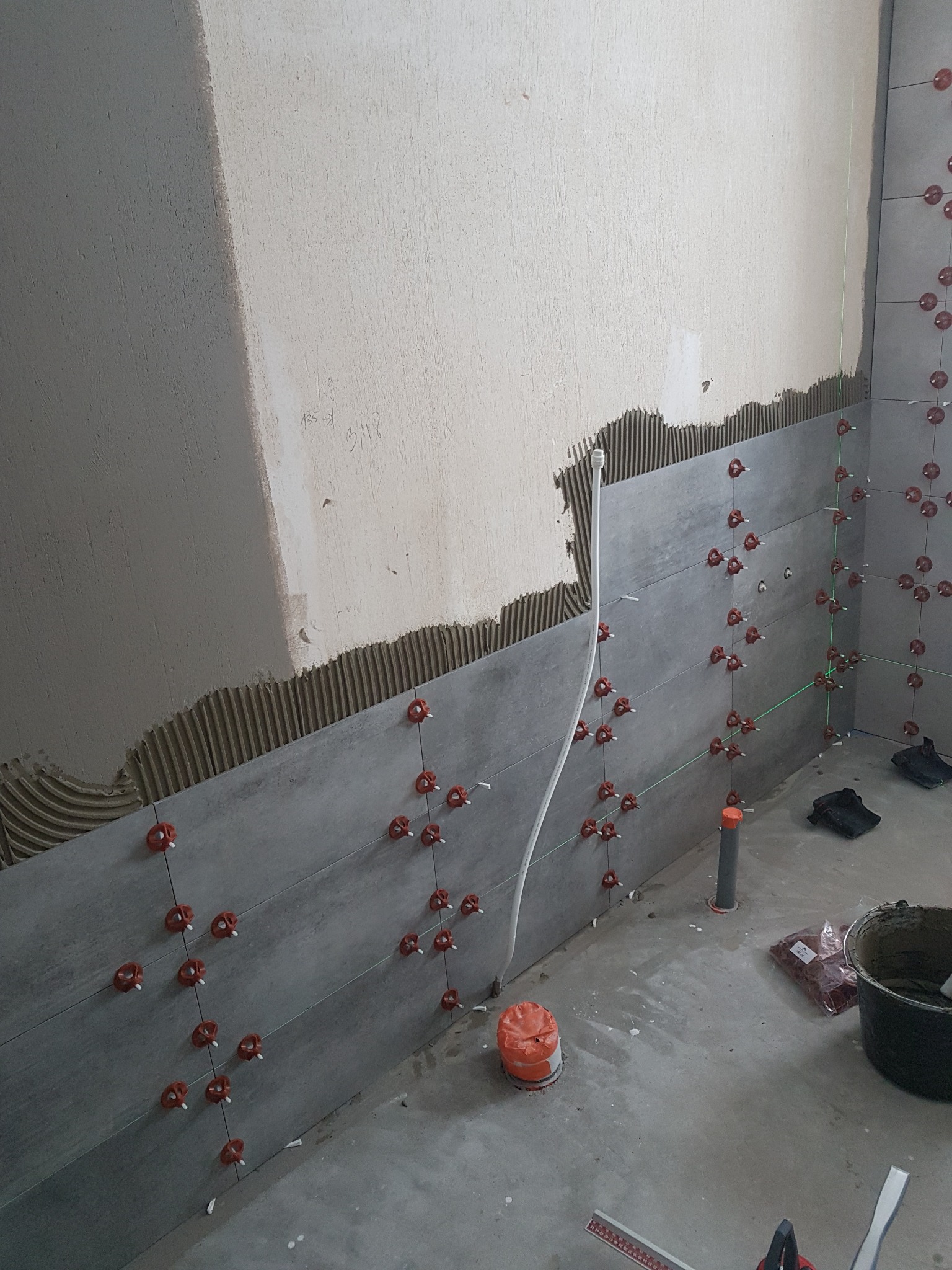 Ściana łazienki w trakcie układania szarych płytek, widoczne krzyżyki dystansowe, poziomica laserowa i nałożony klej na ścianie.