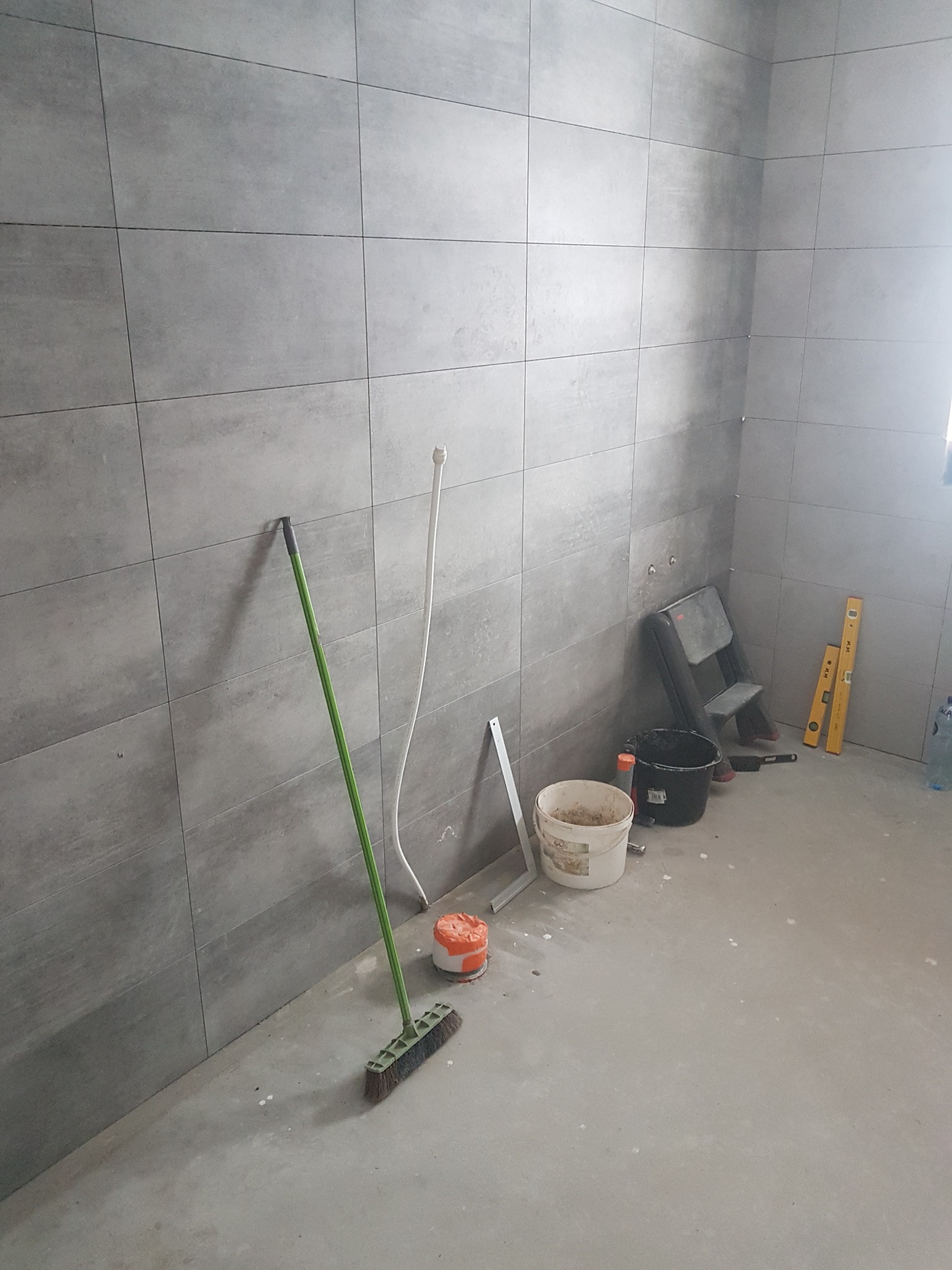 Szara ściana w trakcie układania płytek, widoczne narzędzia: drabina, poziomice, wiadro z klejem, pędzel i rura instalacyjna.