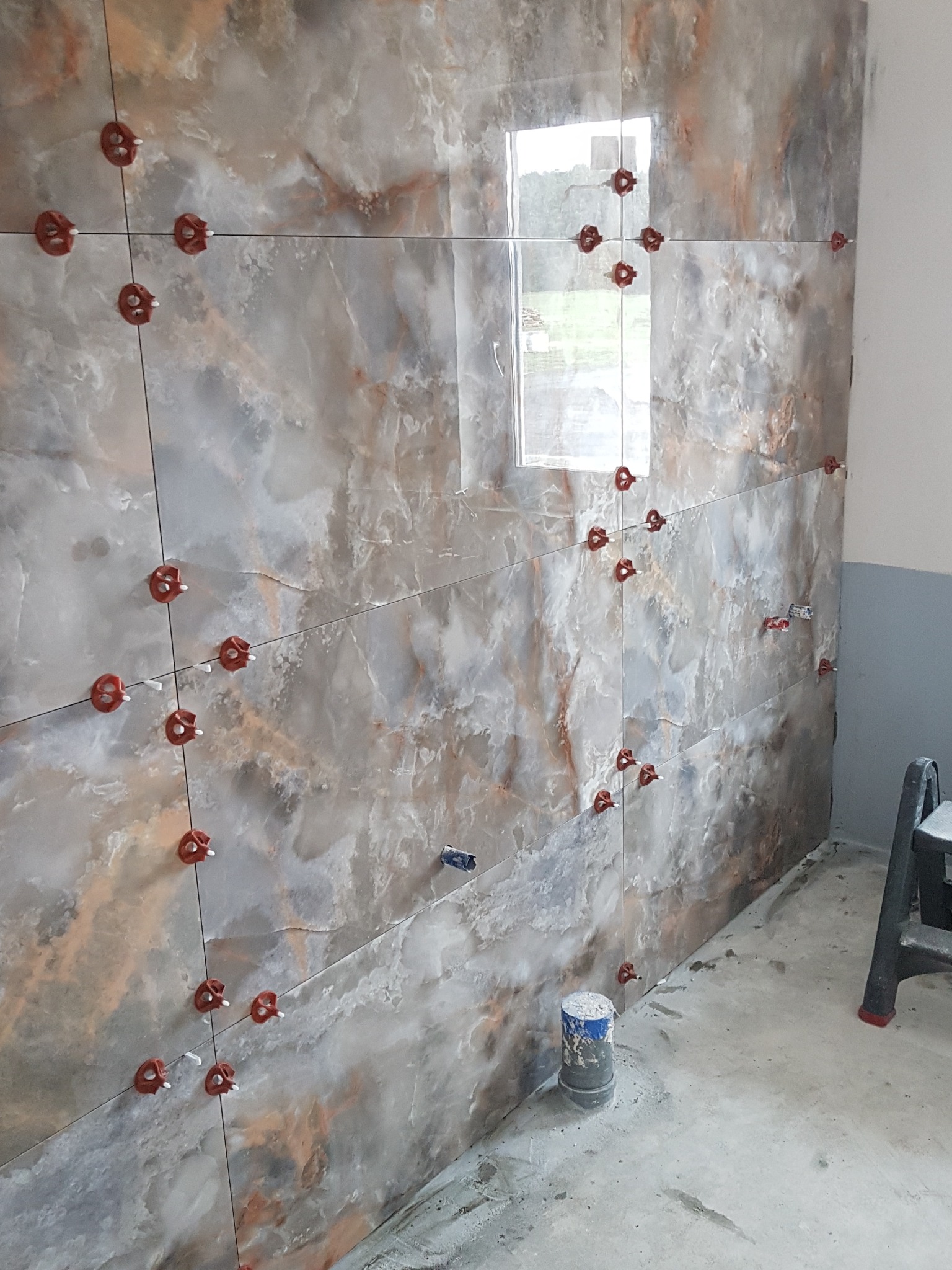 Ściana w trakcie układania płytek imitujących marmur, z użyciem czerwonych klipsów poziomujących, widoczny fragment okna i drabiny.