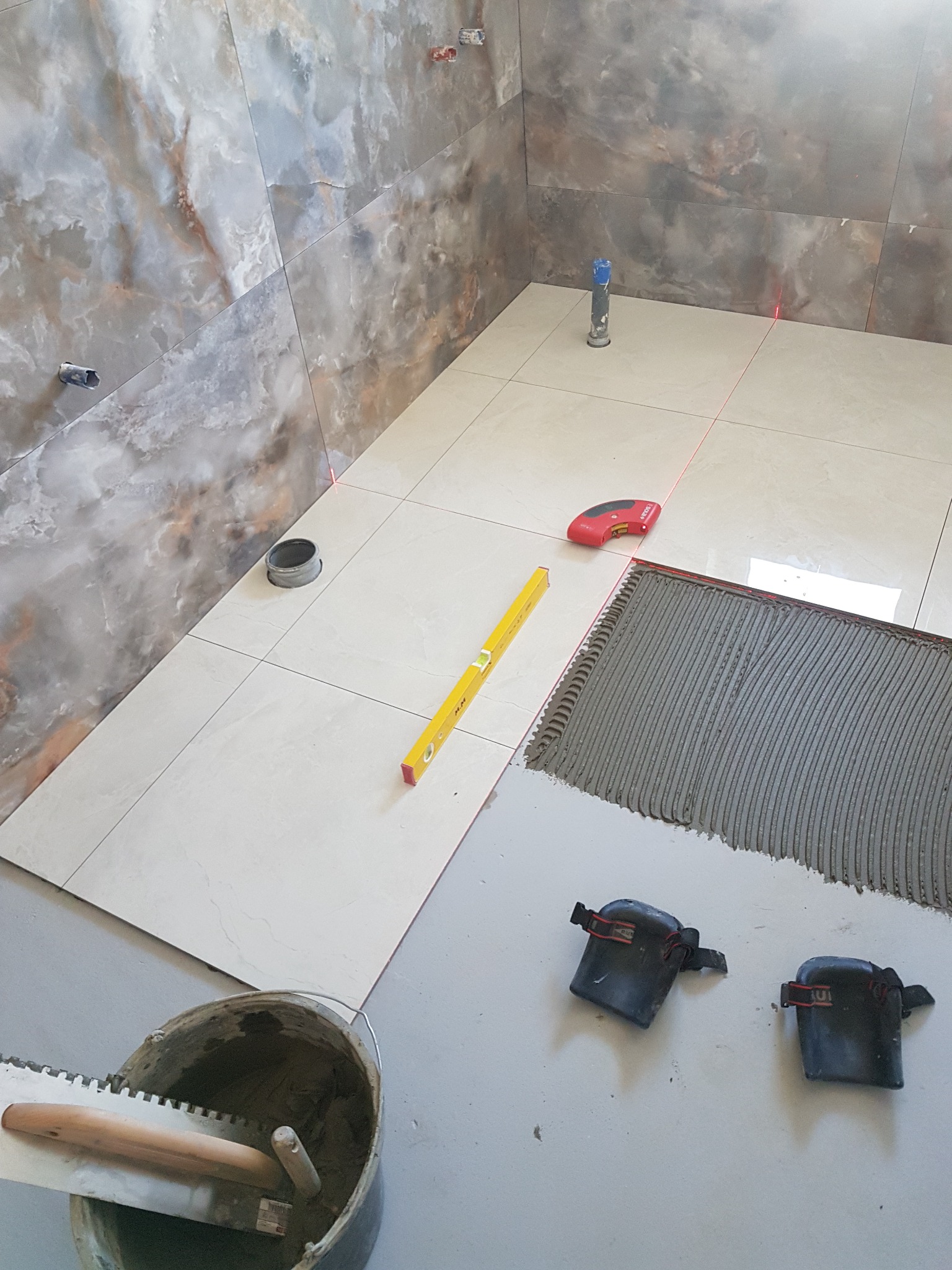 Układanie jasnych płytek podłogowych w łazience z użyciem poziomicy laserowej, kielni z zaprawą i nakolanników.