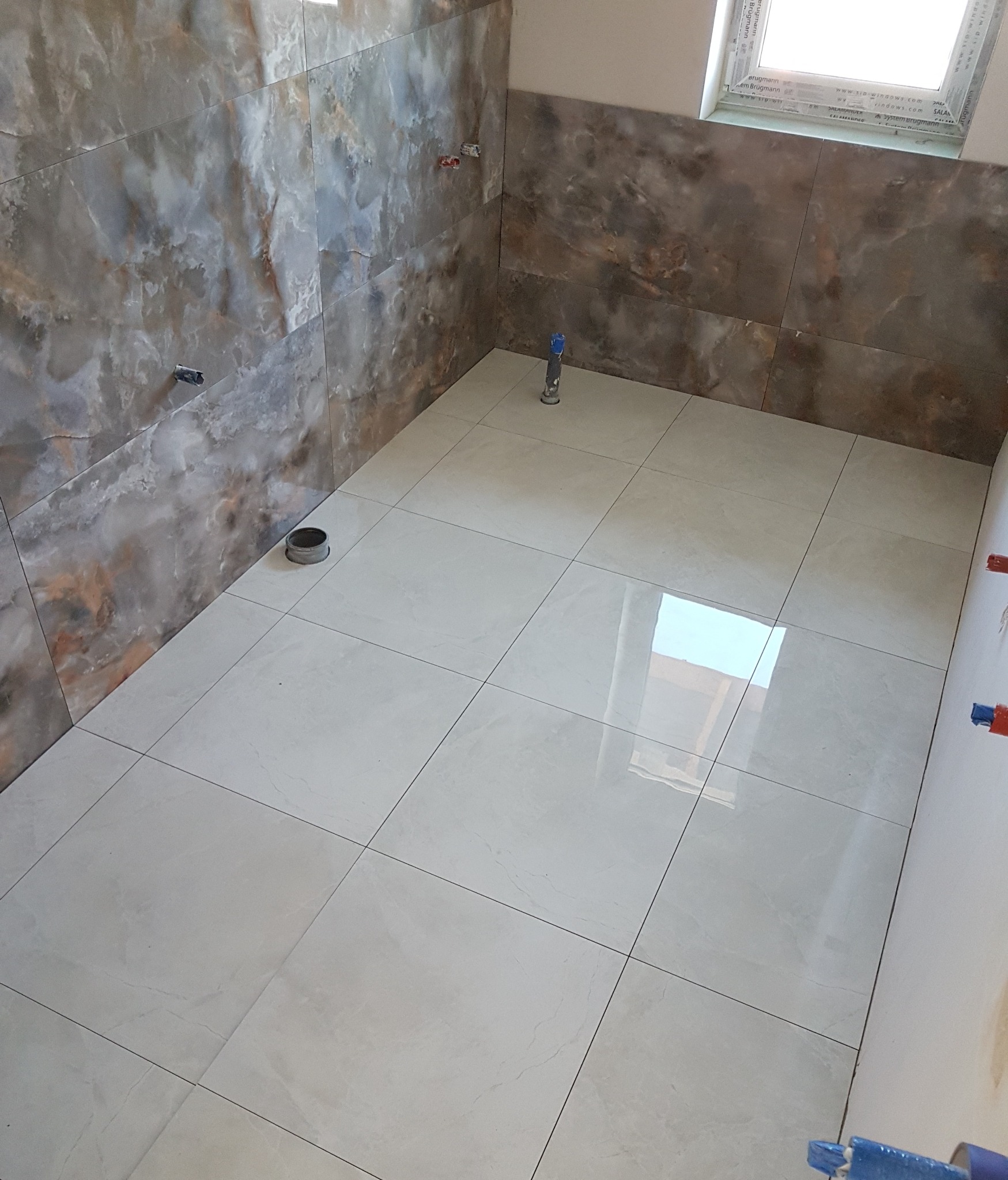 Wykończona łazienka z szarymi marmurkowymi płytkami na ścianach i jasnymi, kwadratowymi płytkami na podłodze. Widoczne wystające rury i otwór kanalizacyjny, okno w górnej części ściany.