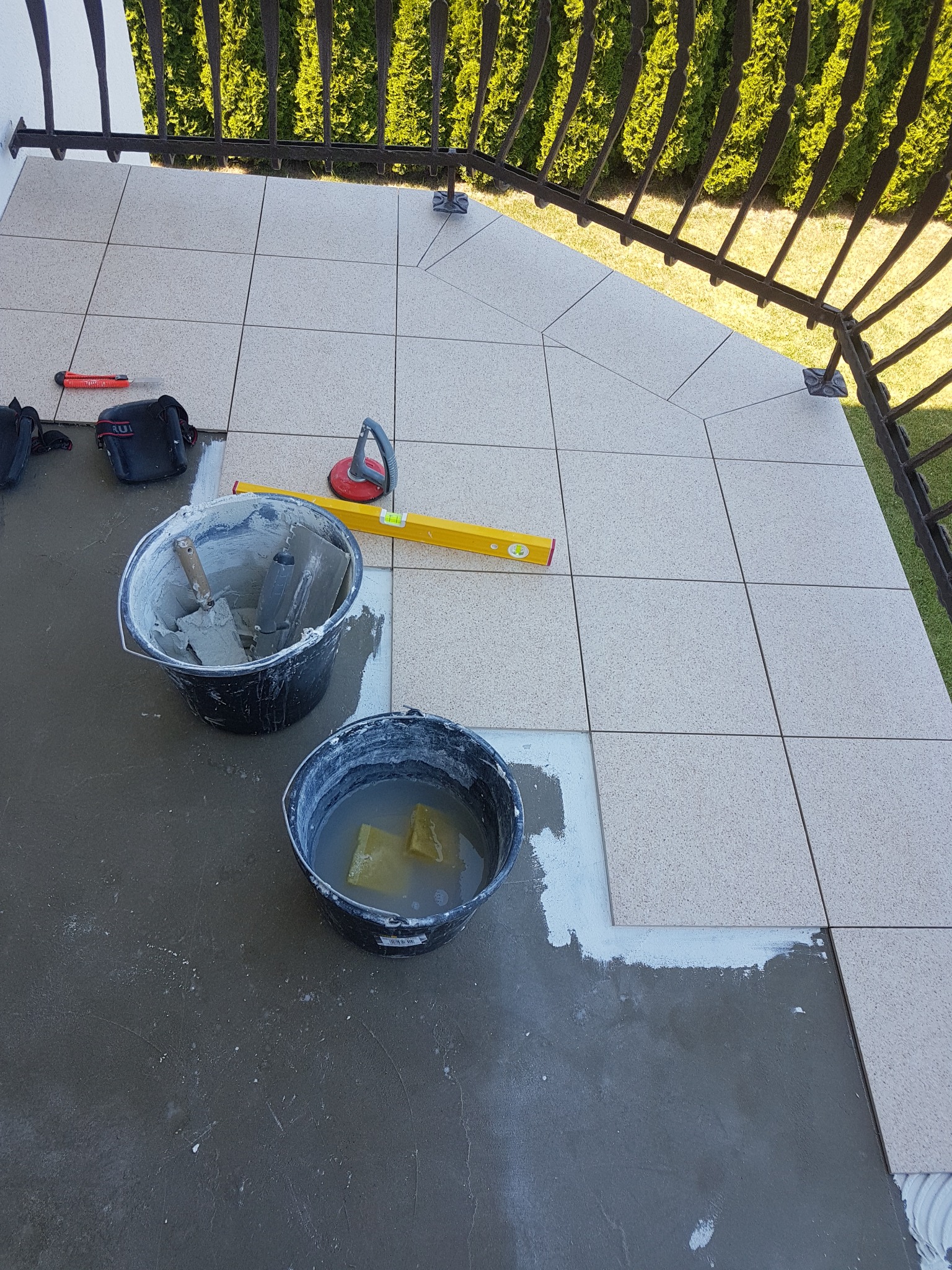 Układanie jasnych płytek na balkonie, widoczne narzędzia: poziomica, wiadro z zaprawą, wiadro z wodą i gąbkami, nóż do cięcia płytek, przyssawka do płytek.