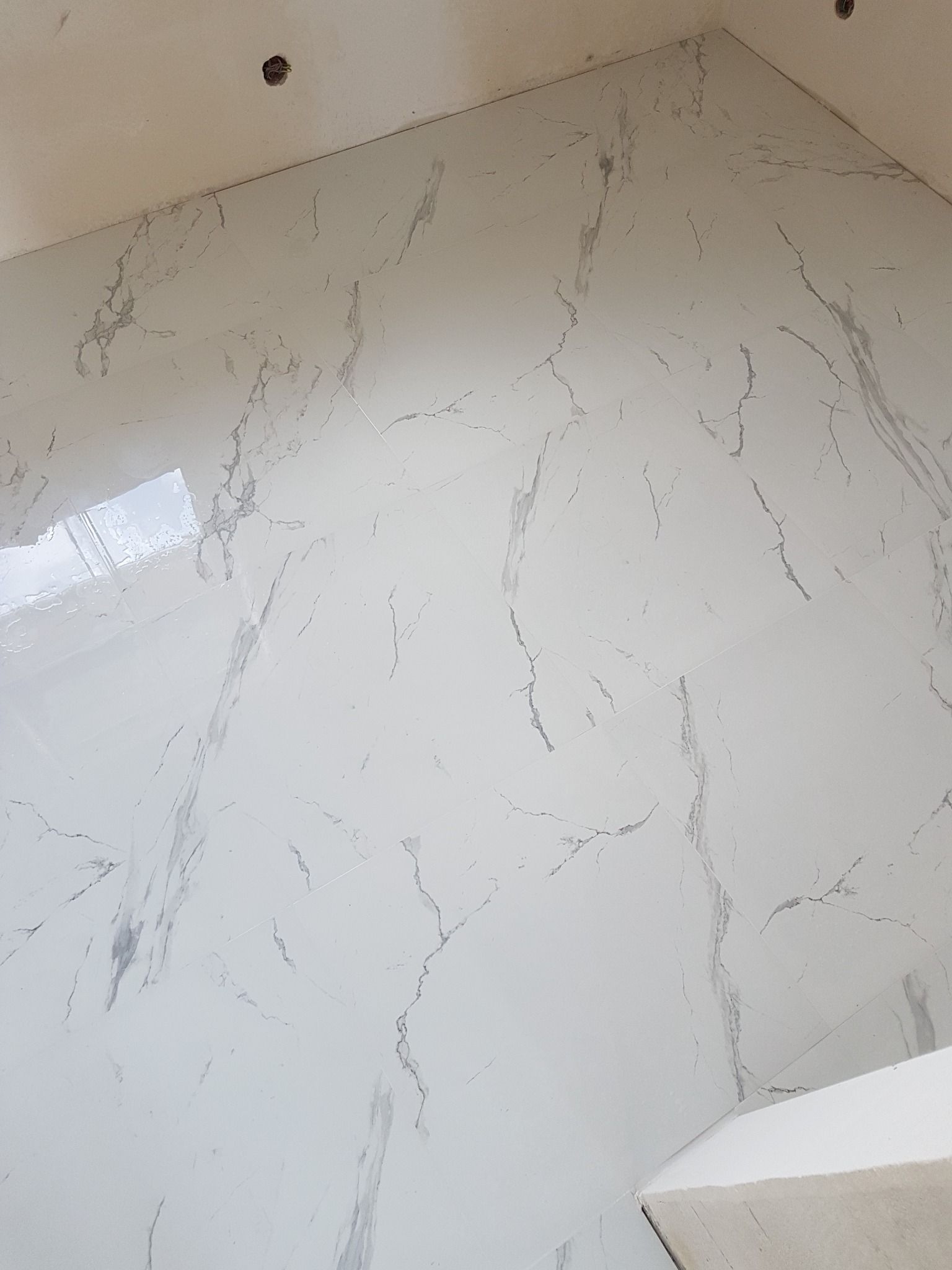 Białe, marmurowe płytki ułożone na ścianie i podłodze w rogu pomieszczenia, widoczne fugi i otwór na instalację elektryczną w suficie.