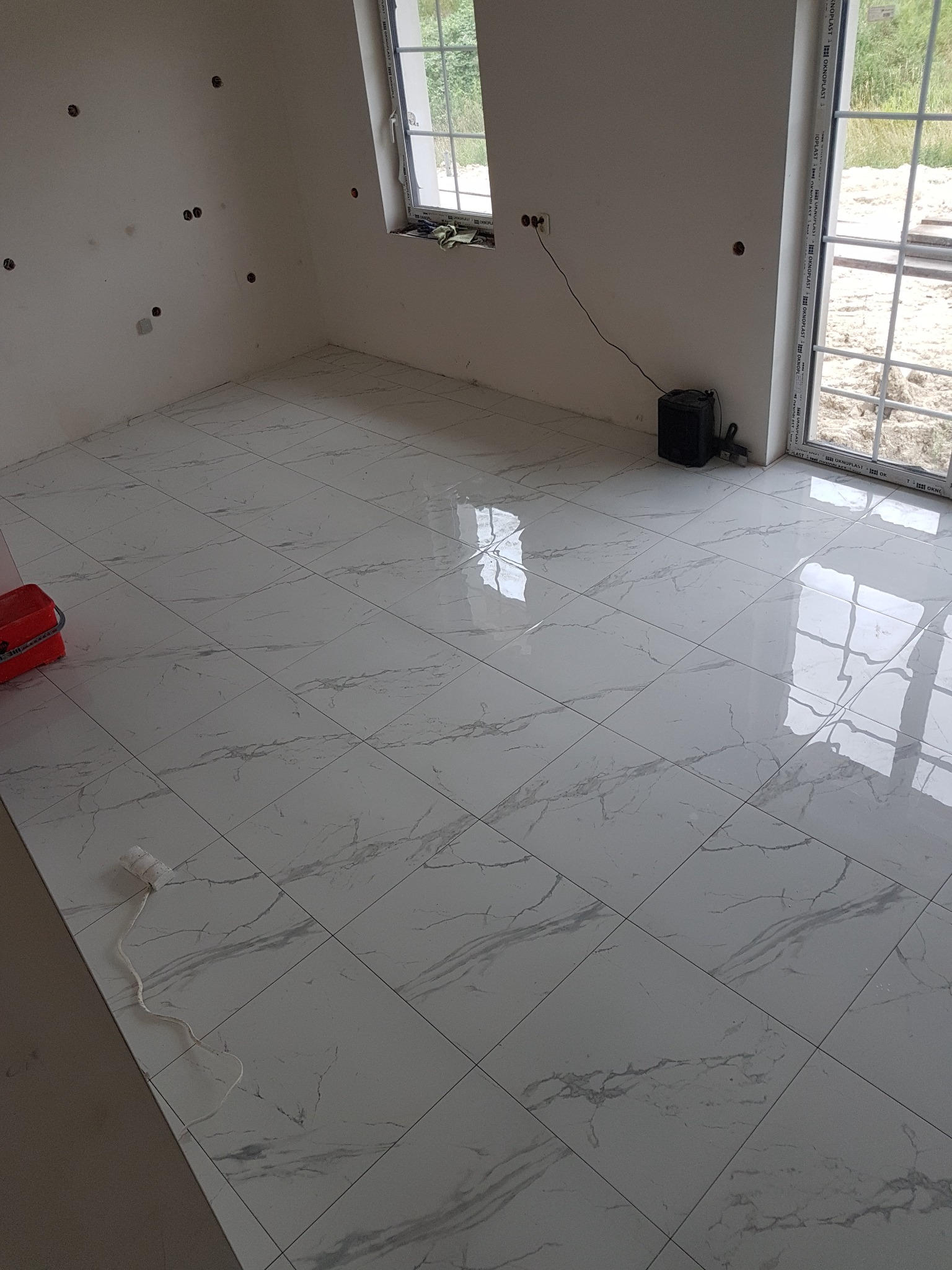 Ułożona, błyszcząca, biała podłoga z płytek imitujących marmur w pustym pomieszczeniu z oknem i przygotowanymi punktami elektrycznymi na ścianie.