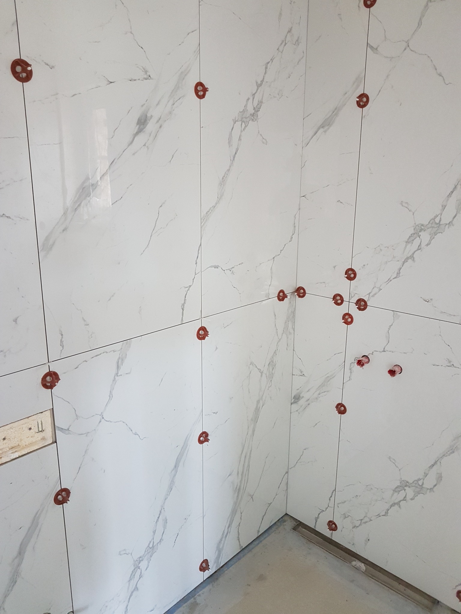 Świeżo ułożone, białe płytki imitujące marmur na ścianach łazienki, z czerwonymi klipsami i sznurkami poziomującymi, widoczne narożniki i fragment odpływu liniowego w podłodze.