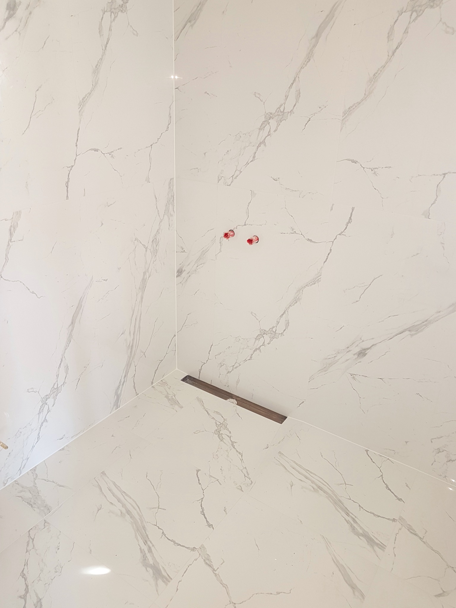 Wykończony narożnik łazienki z białymi płytkami imitującymi marmur, widoczny odpływ liniowy i wystające rury instalacji wodnej.