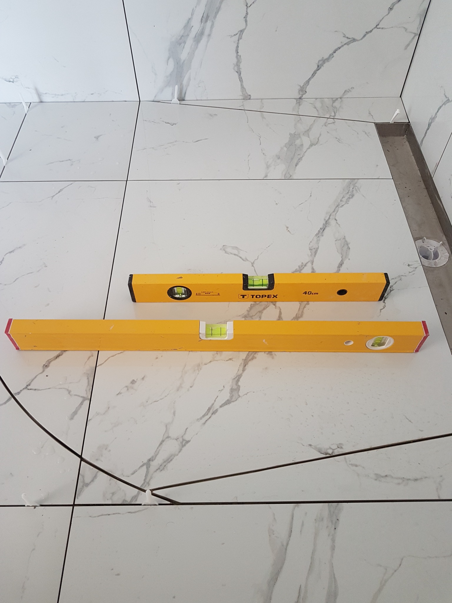 Ułożone białe płytki imitujące marmur, poziomowane dwoma żółtymi poziomicami Topex, widoczne krzyżyki dystansowe i odpływ liniowy.