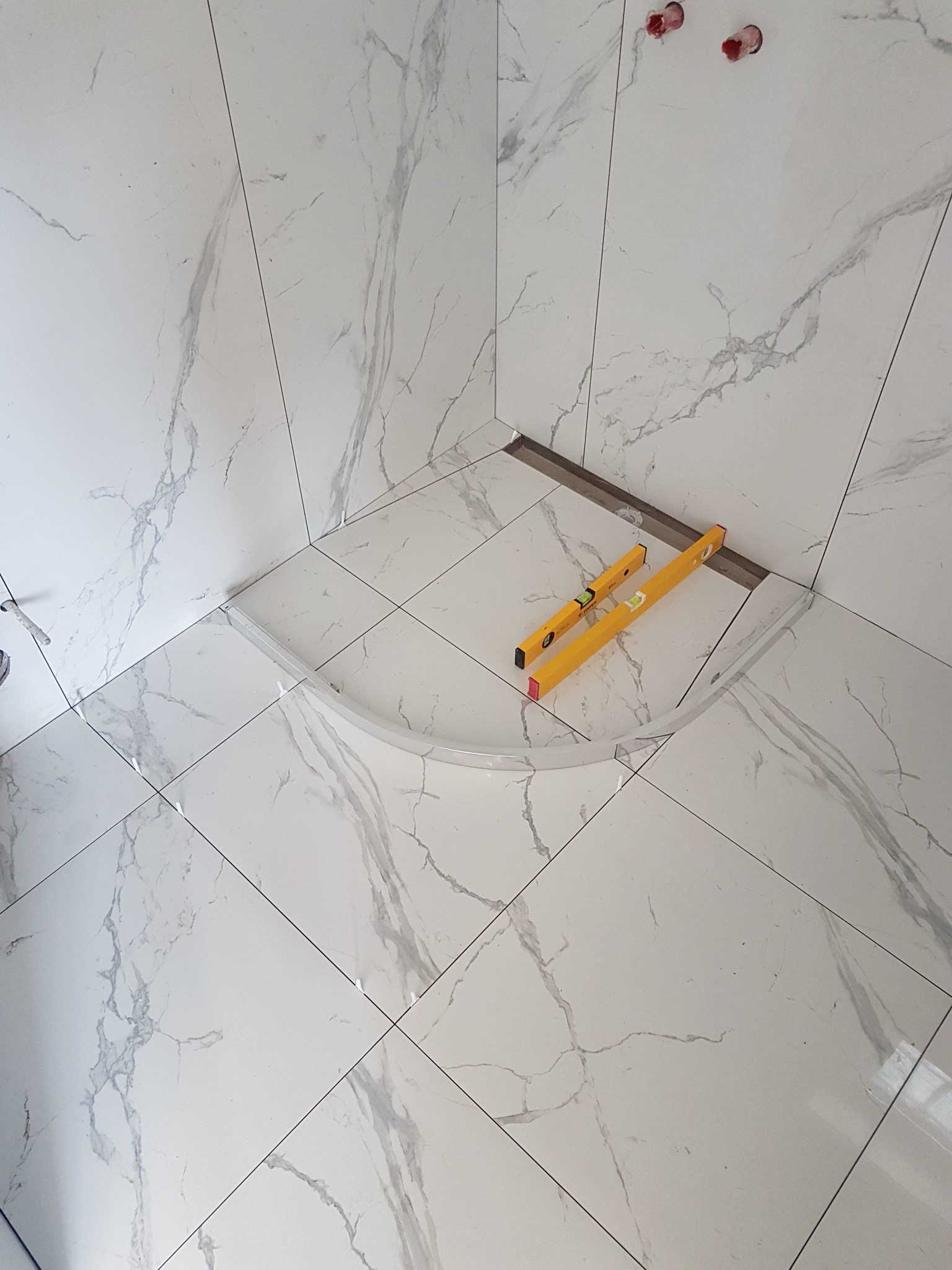 Ułożone białe płytki imitujące marmur w rogu łazienki z wyprofilowanym odpływem liniowym i dwiema żółtymi poziomicami.