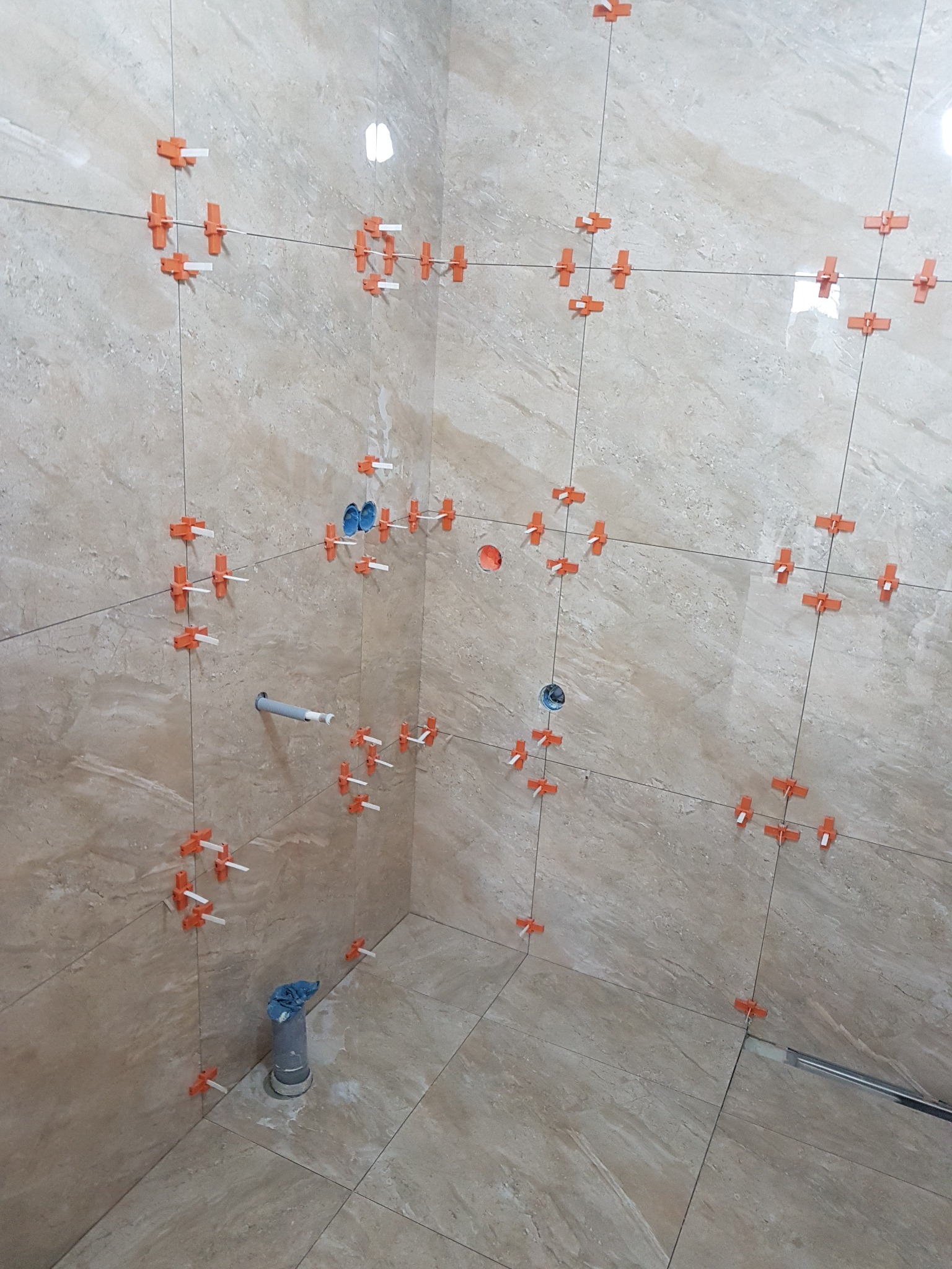 Ściana i podłoga w trakcie układania płytek, widoczne krzyżyki dystansowe i system poziomowania, szare płytki imitujące marmur, przygotowane otwory na instalacje.