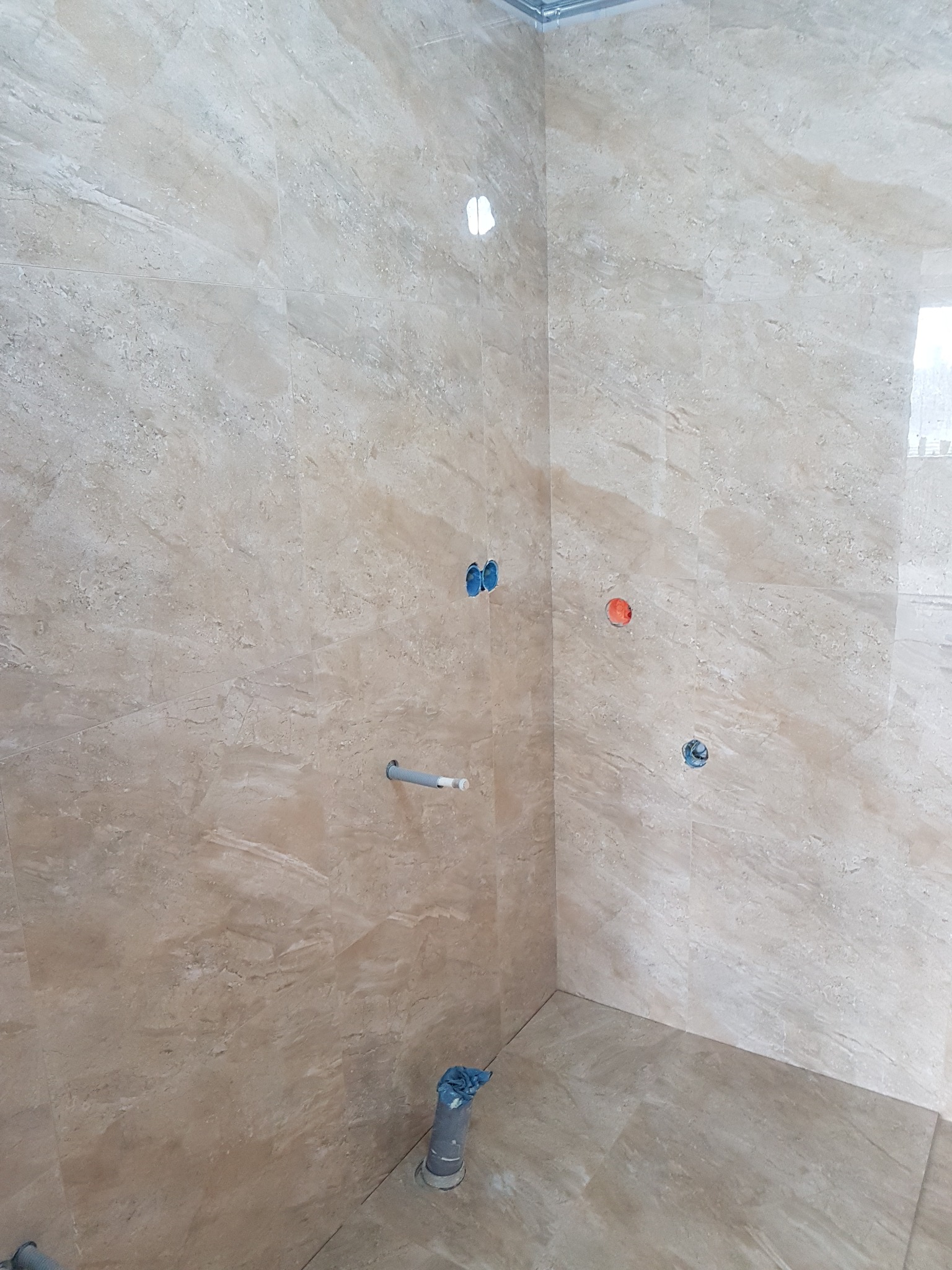 Ściany i podłoga wyłożone beżowymi płytkami imitującymi marmur, widoczne otwory na instalacje, fragment rury kanalizacyjnej zabezpieczonej folią.