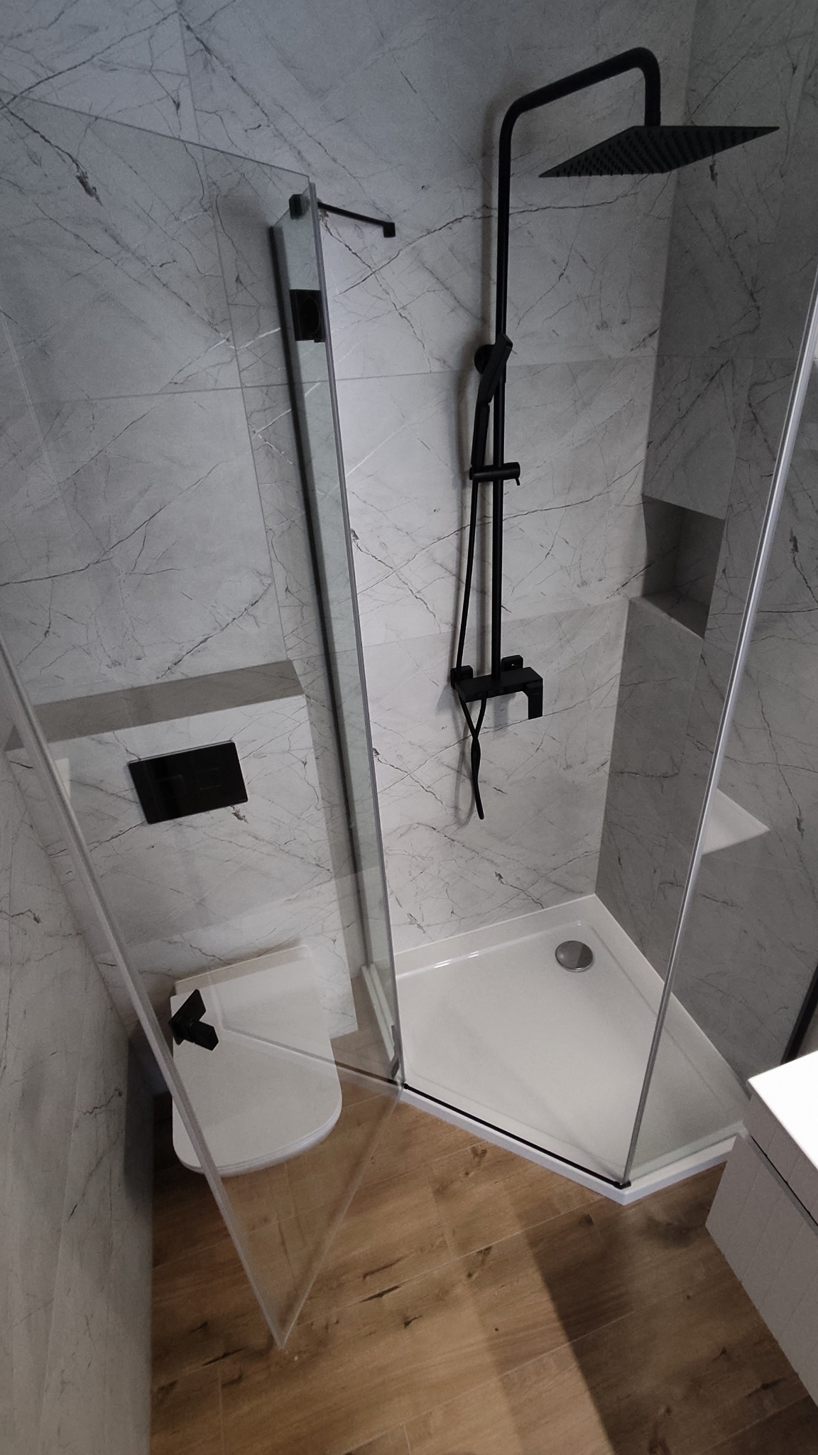 Nowoczesna łazienka z kabiną prysznicową typu walk-in, armaturą w kolorze czarnym i marmurowymi płytkami na ścianach, drewnopodobna podłoga.