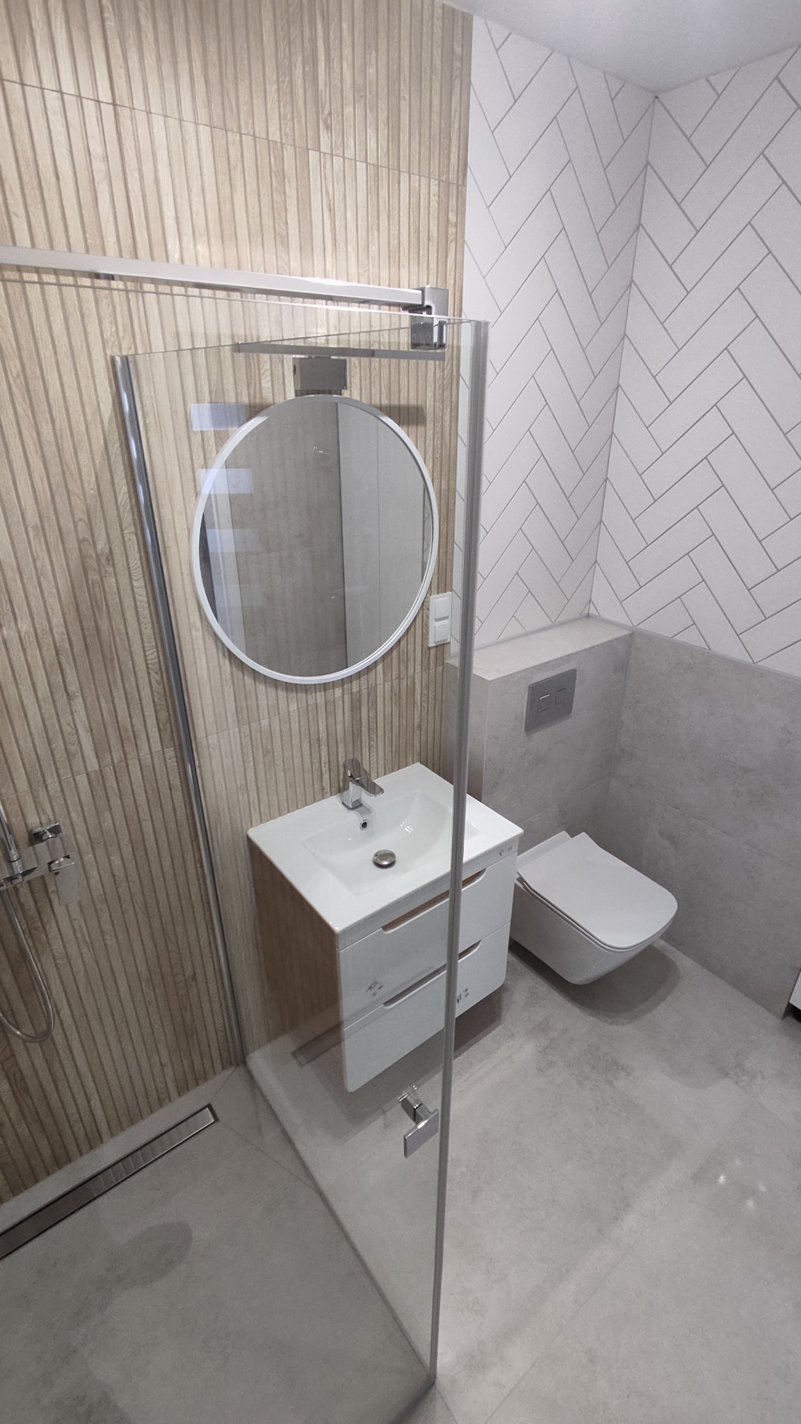 Nowoczesna łazienka z kabiną prysznicową typu walk-in, umywalką z szafką, okrągłym lustrem, toaletą podwieszaną i płytkami imitującymi drewno na ścianie.