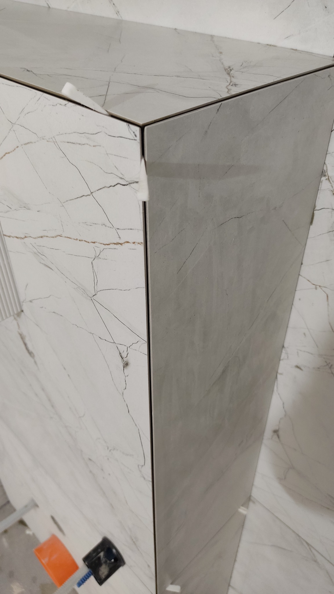 Fragment narożnika obudowanego płytkami imitującymi marmur z widocznymi narzędziami glazurniczymi w dolnej części kadru.