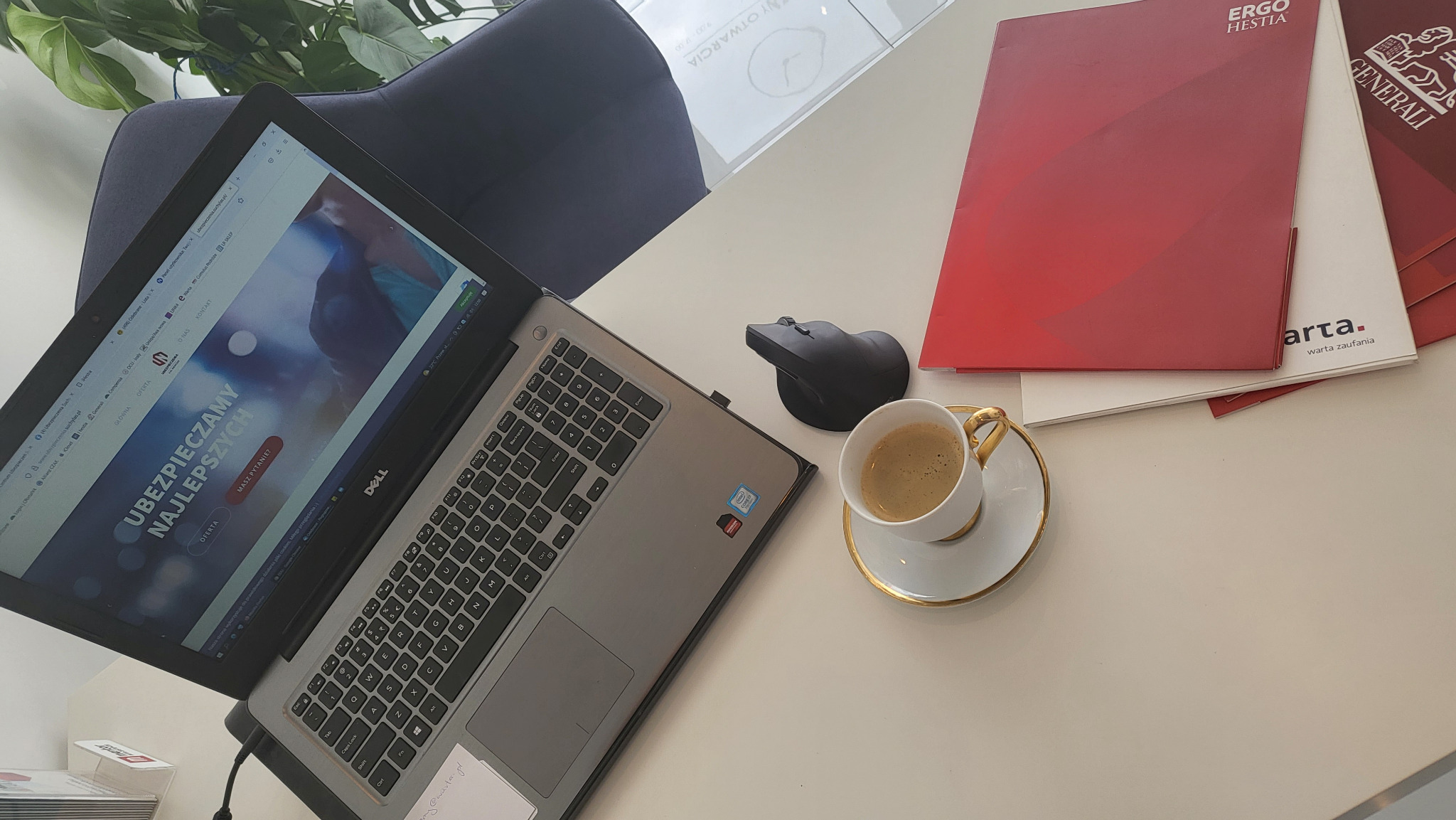 Laptop z otwartą stroną internetową o ubezpieczeniach, obok myszka ergonomiczna, filiżanka kawy i teczki z logotypami firm ubezpieczeniowych Ergo Hestia i Generali na białym biurku.