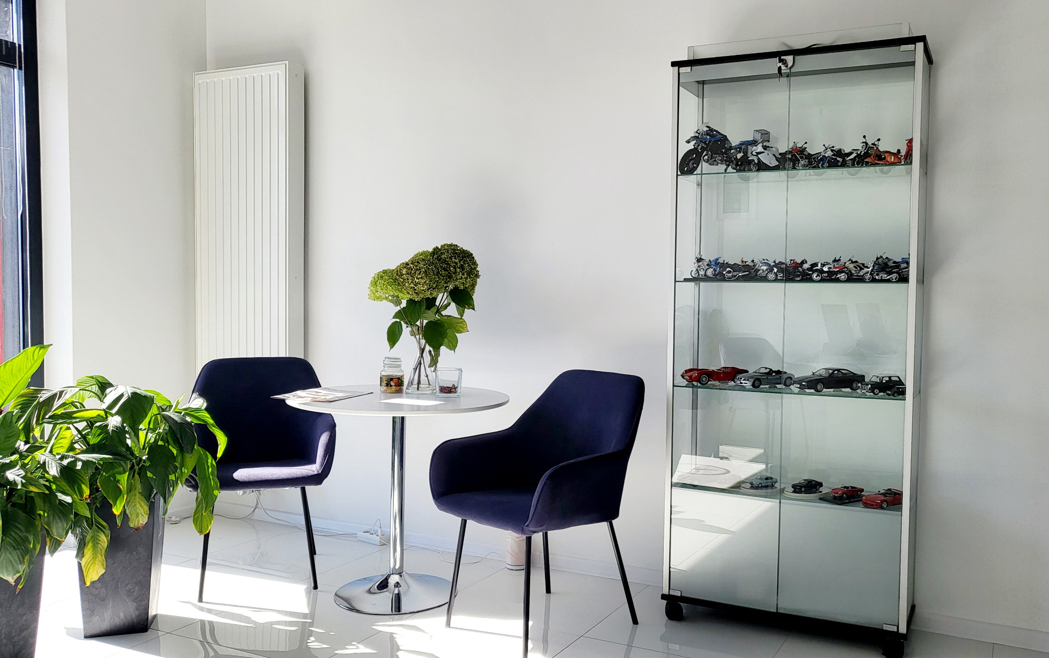 Jasne wnętrze biura z minimalistycznym stolikiem, krzesłami i przeszkloną witryną wypełnioną modelami motocykli i samochodów.