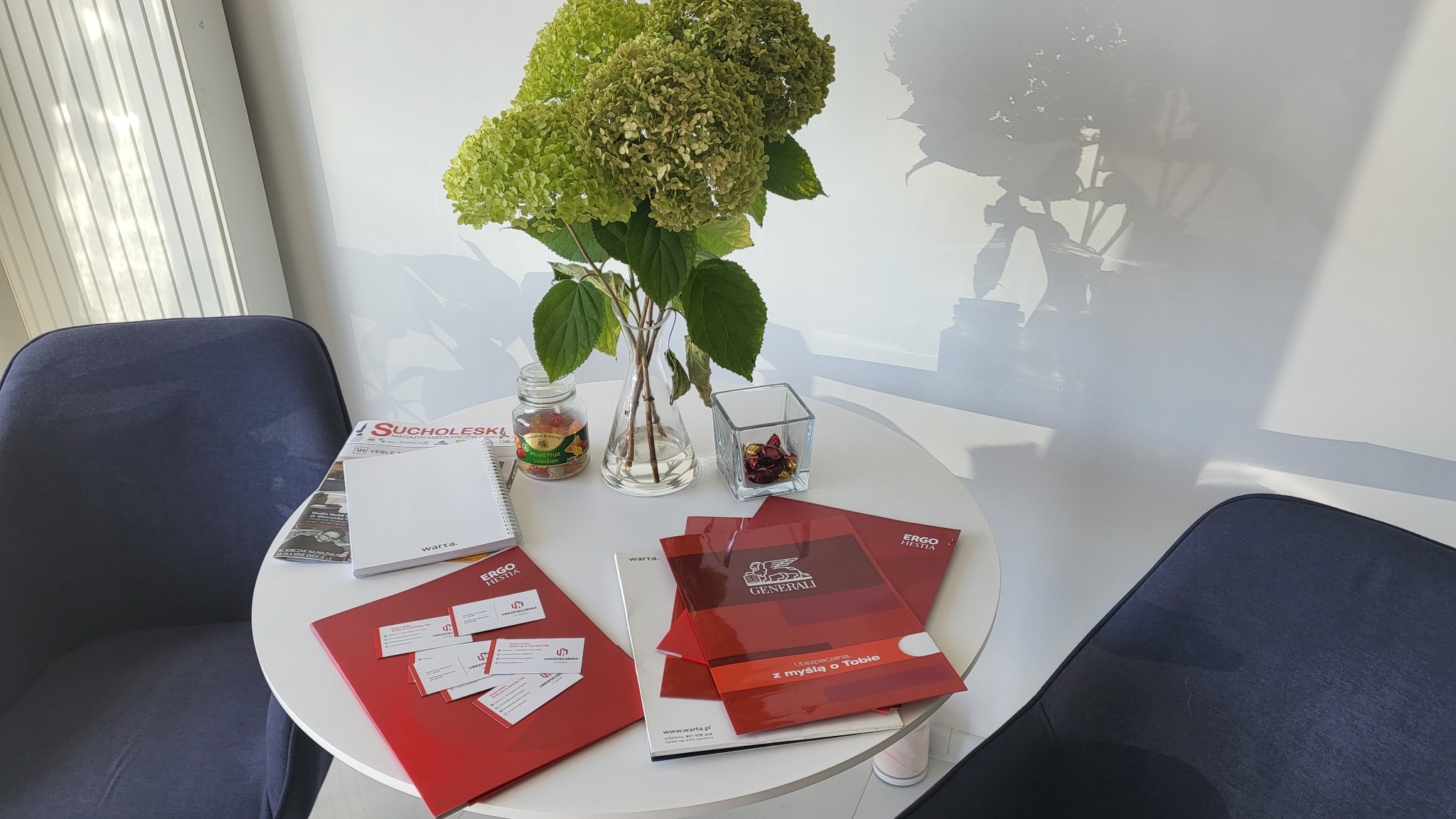 Stół w biurze z materiałami informacyjnymi firm ubezpieczeniowych Ergo Hestia, Warta i Generali, wazonem z hortensjami i dwoma krzesłami w tle