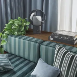 Stylowa aranżacja wnętrza z zasłonami w paski, roletami rzymskimi w tle, zielonymi poduszkami w paski, rośliną doniczkową i dekoracyjną lampą na drewnianym blacie.