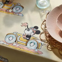 Beżowy obrus z nadrukiem Myszki Miki w samochodach wyścigowych, szklanka z wodą i różowy talerz na plecionym podkładce.