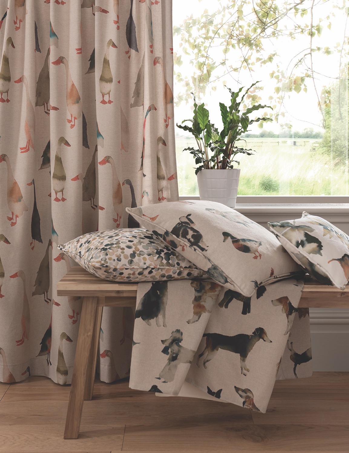 Beżowe zasłony z motywem kaczek, poduszki z różnymi wzorami zwierząt leżące na drewnianej ławce, roślina doniczkowa na parapecie, widok na zielone pole za oknem.