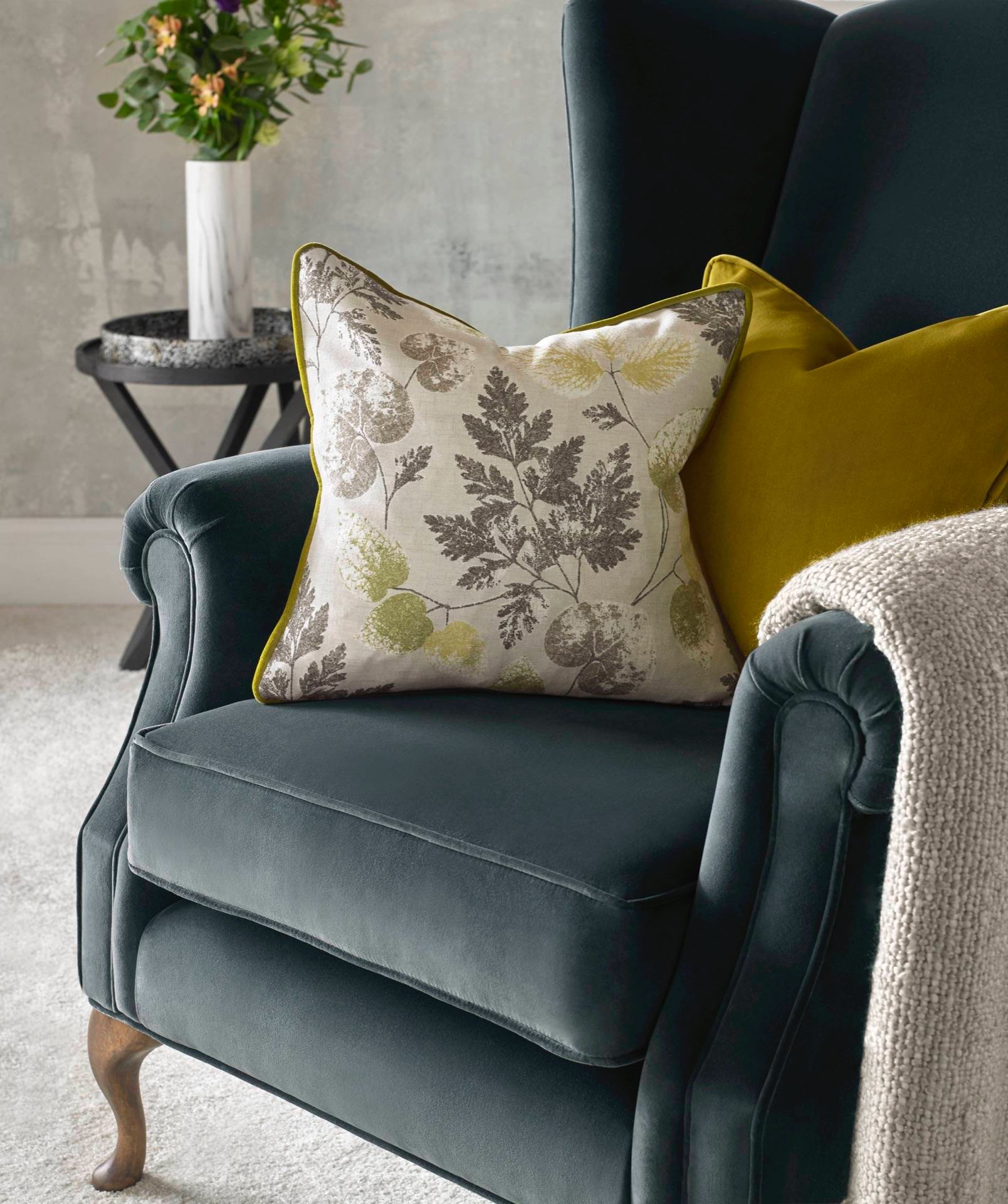 Granatowy fotel typu uszak z dekoracyjną poduszką w liście i żółtą lamówką, obok żółta poduszka i beżowy pled, w tle stolik z kwiatami.