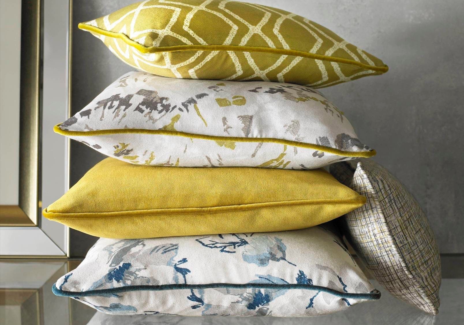 Stos czterech dekoracyjnych poduszek: geometryczny wzór w odcieniach żółci, kwiatowy motyw w szarościach i żółciach, jednolita żółta, oraz kwiatowy motyw w błękitach i szarościach, każda z lamówką...