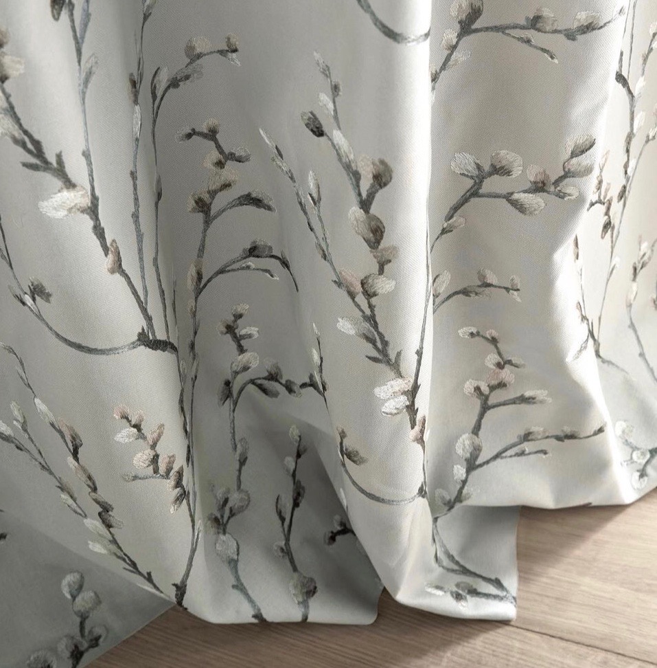Jasnoszara tkanina zasłonowa z motywem gałązek kwitnącej wierzby, ułożona w dekoracyjne fałdy, widoczny fragment drewnianej podłogi.