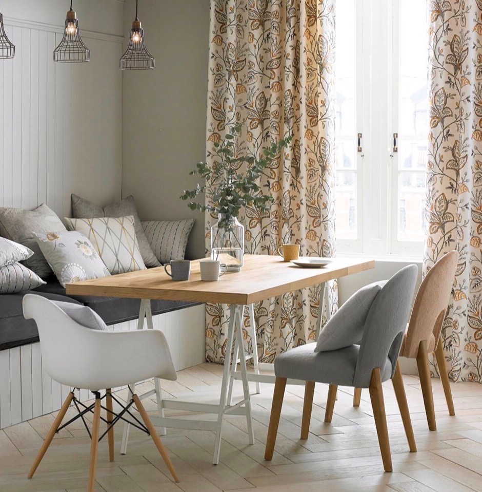 Jasne, przestronne wnętrze jadalni w Krakowie z zasłonami w roślinny wzór, drewniany stół z filiżankami i wazonem, oraz różnokolorowe krzesła, oświetlone wiszącymi lampami.