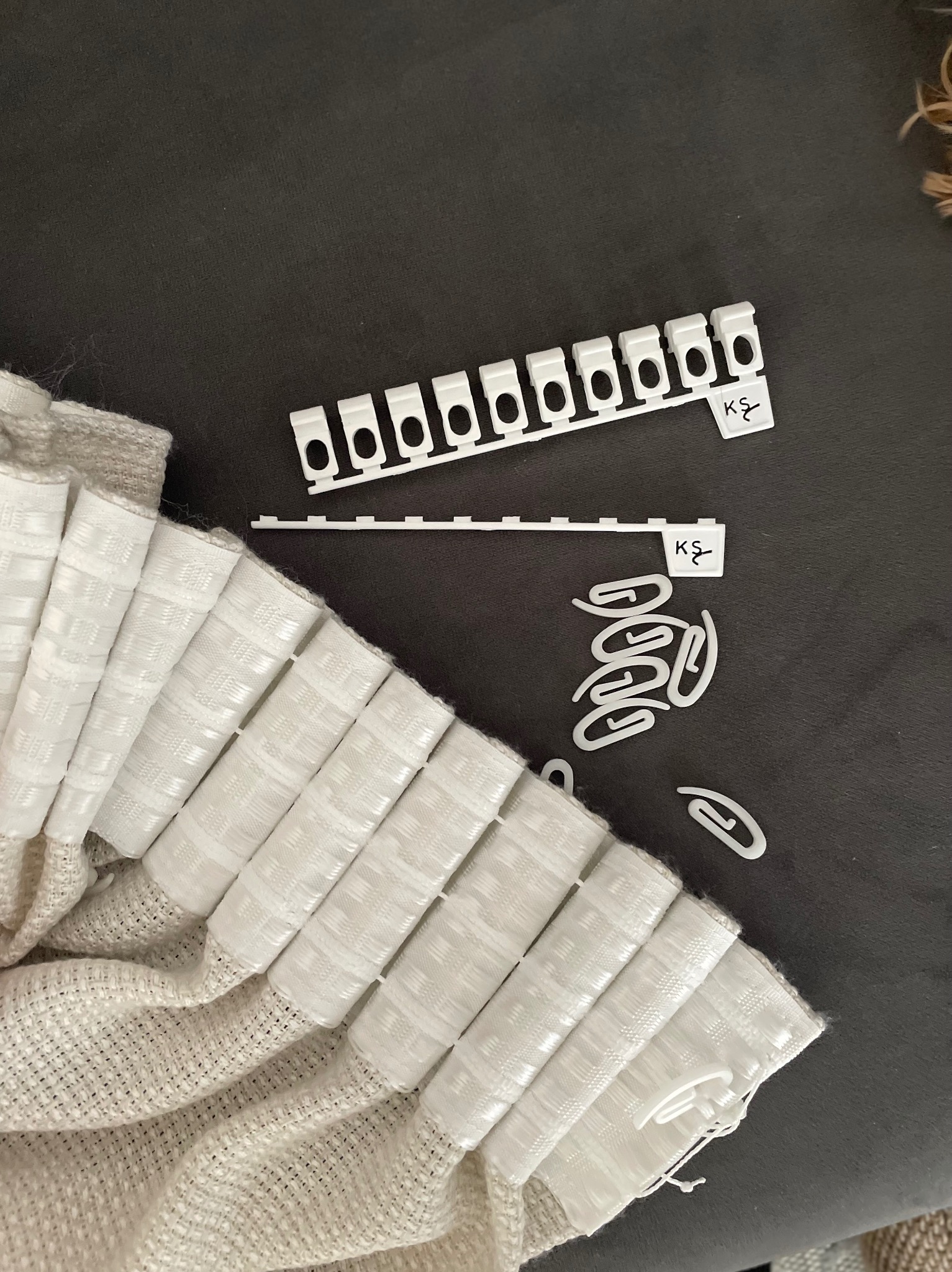 Fragment marszczonej, beżowej tkaniny z wszytymi, białymi taśmami, obok listwa i żabki do zawieszania na karniszu.