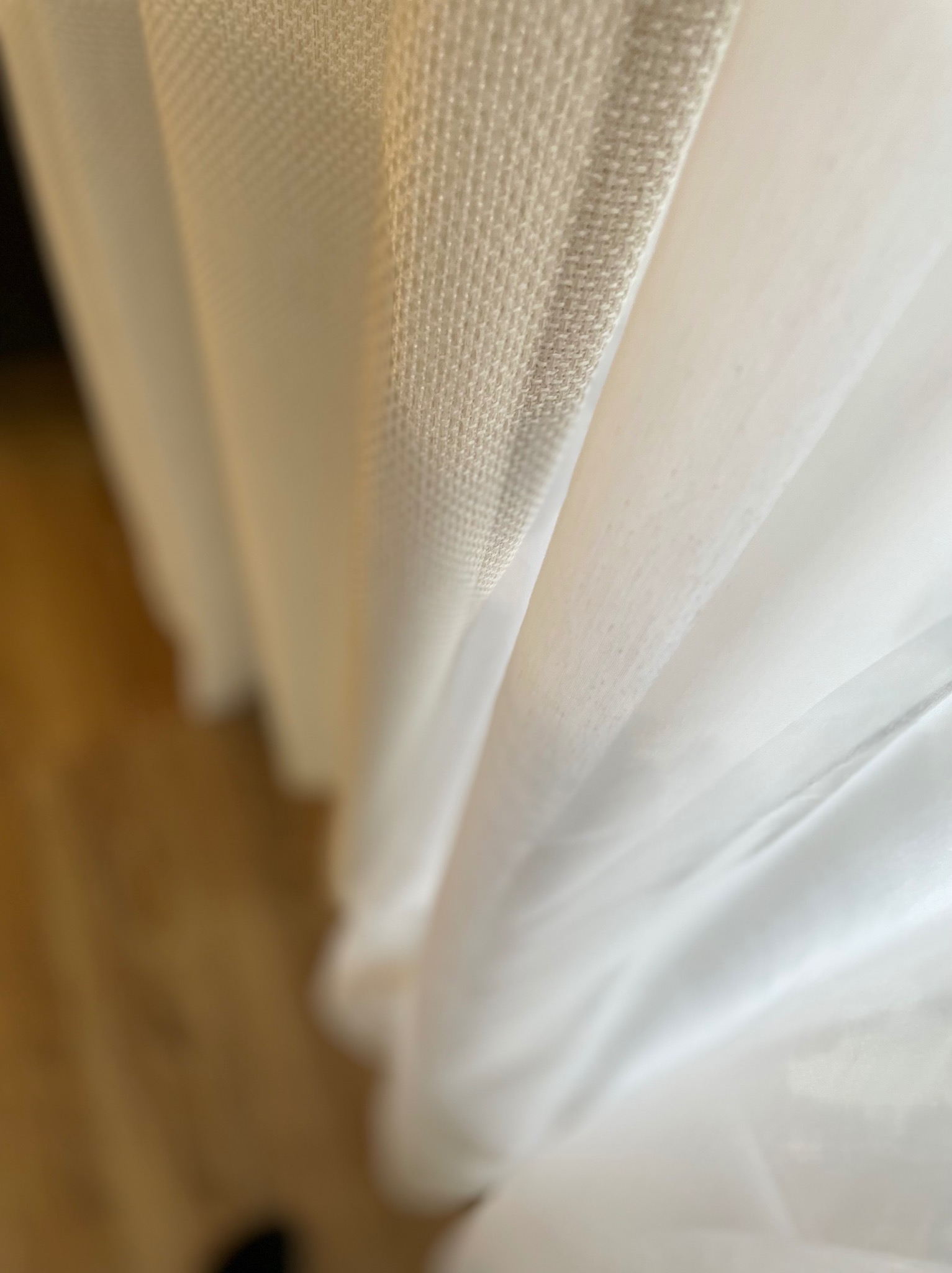 Detale delikatnej, białej, prześwitującej firany z beżową, grubszą tkaniną u góry, z widocznym fragmentem drewnianej podłogi w tle.