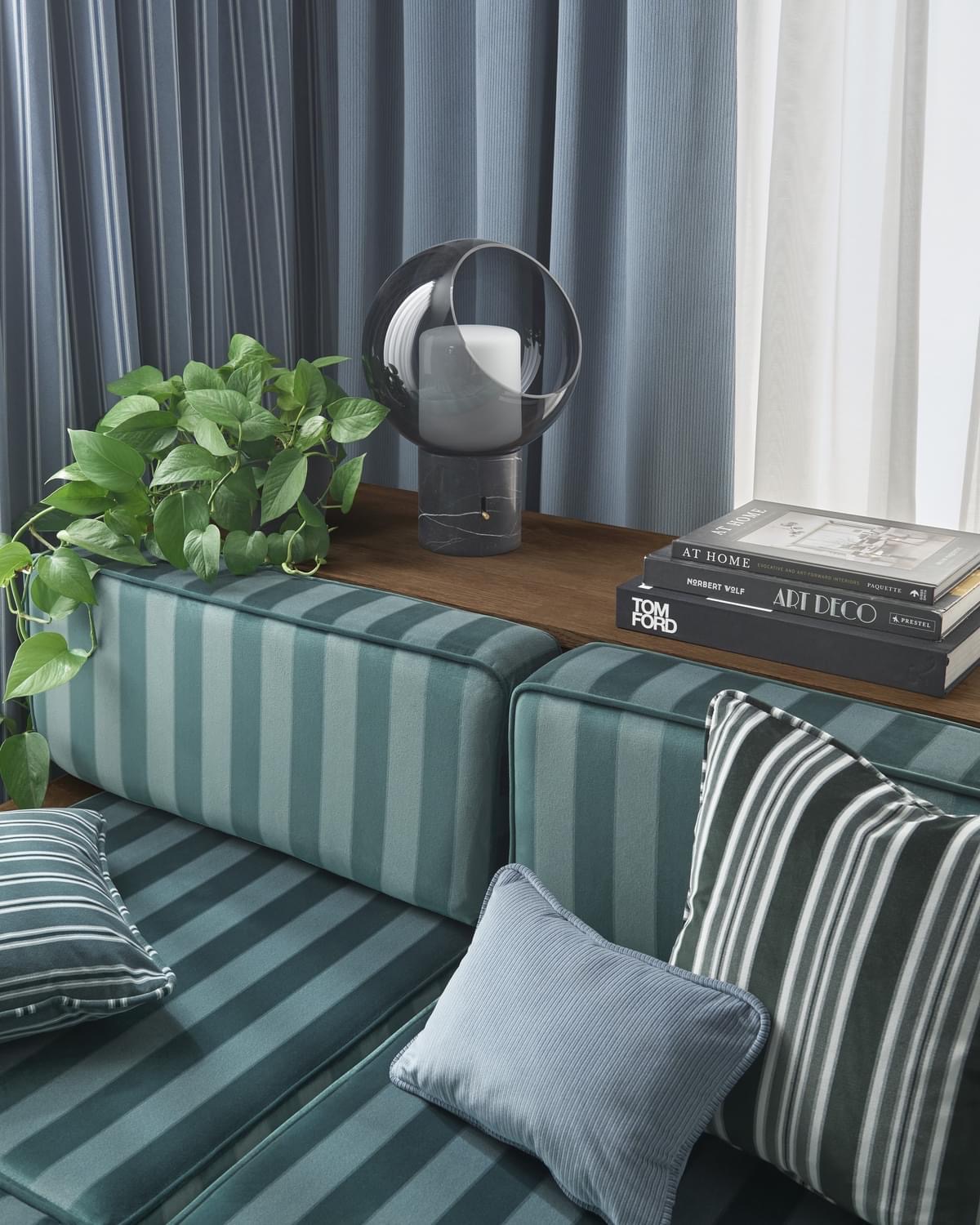 Stylowa aranżacja wnętrza z zasłonami w paski, roletami rzymskimi w tle, zielonymi poduszkami w paski, rośliną doniczkową i dekoracyjną lampą na drewnianym blacie.