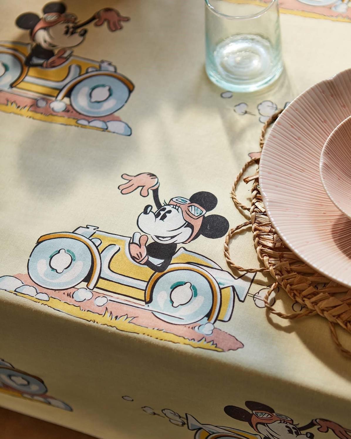 Beżowy obrus z nadrukiem Myszki Miki w samochodach wyścigowych, szklanka z wodą i różowy talerz na plecionym podkładce.